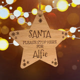 A wooden star-shaped hanging ornament engraved with 'SANTA PLEASE STOP HERE FOR Alfie' and small snowflake details, shown against a warm bokeh background with string lights.