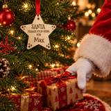 A wooden star-shaped Christmas ornament hanging on a decorated tree, with a person in a Santa hat and white glove reaching for a wrapped gift underneath. The ornament reads 'SANTA PLEASE STOP HERE FOR Max'.