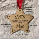 A star-shaped wooden Christmas ornament engraved with 'SANTA PLEASE STOP HERE FOR Max' and small snowflake designs, hanging from a red ribbon on a textured white faux fur background. Text overlays provide product details including size, material, and customization options.
