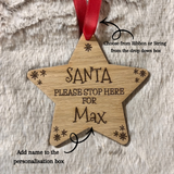A star-shaped wooden Christmas ornament engraved with 'SANTA PLEASE STOP HERE FOR Max' and small snowflake details, hanging from a red ribbon on a faux fur background with instructional text overlays.
