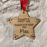 A star-shaped wooden Christmas ornament engraved with 'SANTA PLEASE STOP HERE FOR Max' and small snowflake designs, hanging from a red ribbon on a textured white faux fur background.