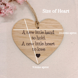 A heart-shaped wooden hanging plaque with engraved text reading 'A new little hand to hold. A new little heart to love' and a small heart cutout near the bottom.