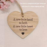 A heart-shaped wooden keepsake plaque engraved with the quote 'A new little hand to hold. A new little heart to love' and a small heart at the bottom, hanging from a jute string on a textured beige fabric background with pink dried flowers in the corner.