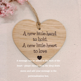 A heart-shaped wooden hanging plaque with a natural wood grain finish, featuring the engraved message 'A new little hand to hold. A new little heart to love' in black text, suspended by a twine loop.