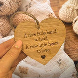 A heart-shaped wooden hanging ornament engraved with the phrase 'A new little hand to hold. A new little heart to love' and held by a hand, set against a soft, cozy background of a teddy bear and knitted baby booties.