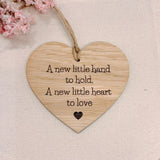 A heart-shaped wooden hanging plaque with the engraved phrase 'A new little hand to hold. A new little heart to love' and a small heart at the bottom, suspended by twine on a light fabric background with pink flowers in the corner.