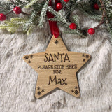 A wooden star-shaped Christmas ornament engraved with 'SANTA PLEASE STOP HERE FOR Max' and small snowflake details, hanging from a red ribbon amidst festive greenery and red berries on a white faux fur surface.